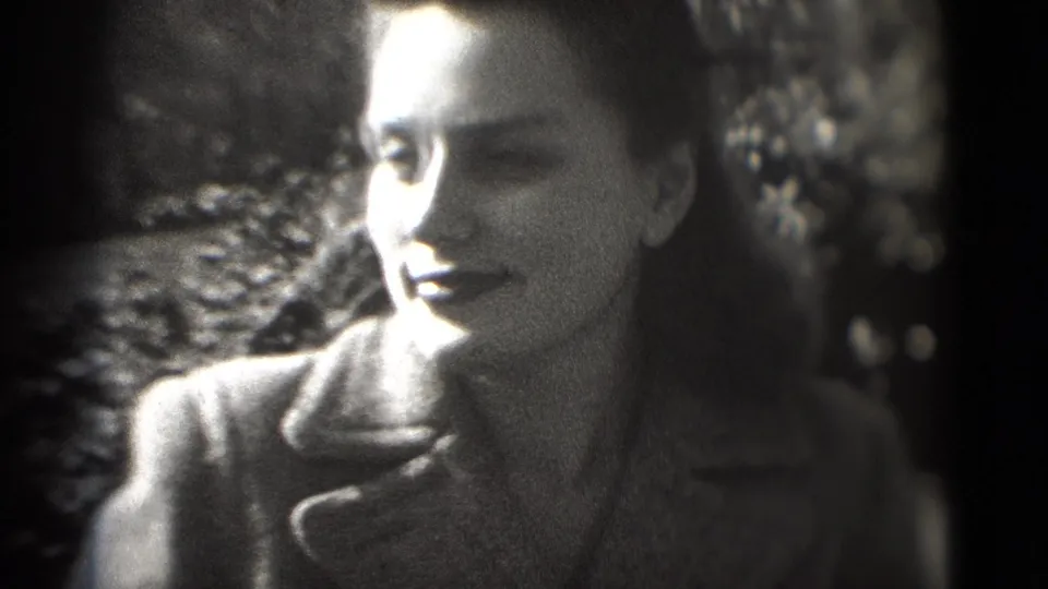 1975 8mm frame shows the woman turning slightly in profile, smiling in a dark coat against a leafy outdoor background.