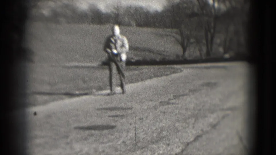 1975 8mm home-movie frame shows a man at a distance on a dirt or gravel roadside with open grass and trees nearby.