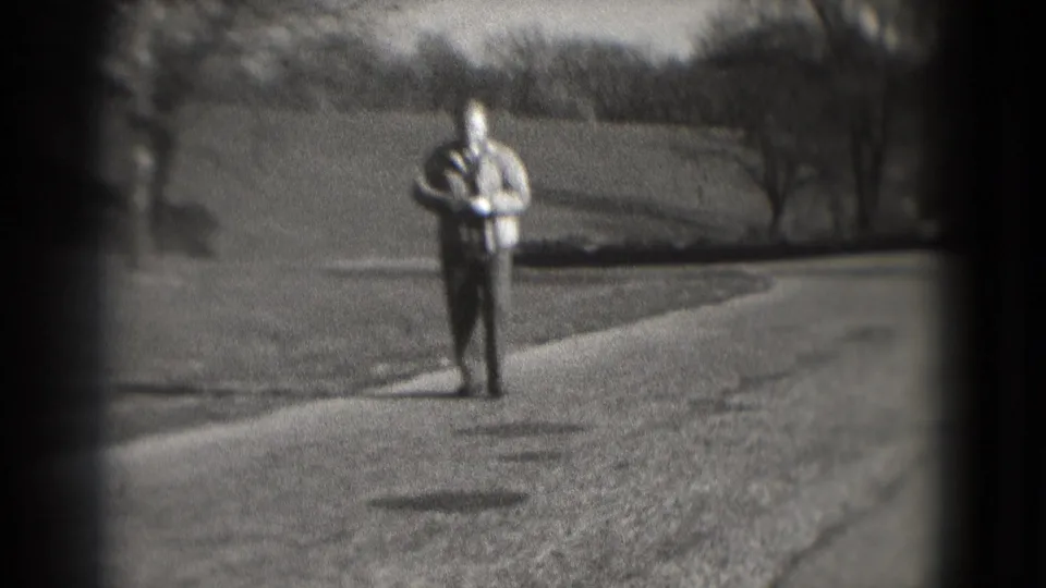 1975 8mm frame shows the man on a dirt or gravel road, with grassy ground and trees in the background.