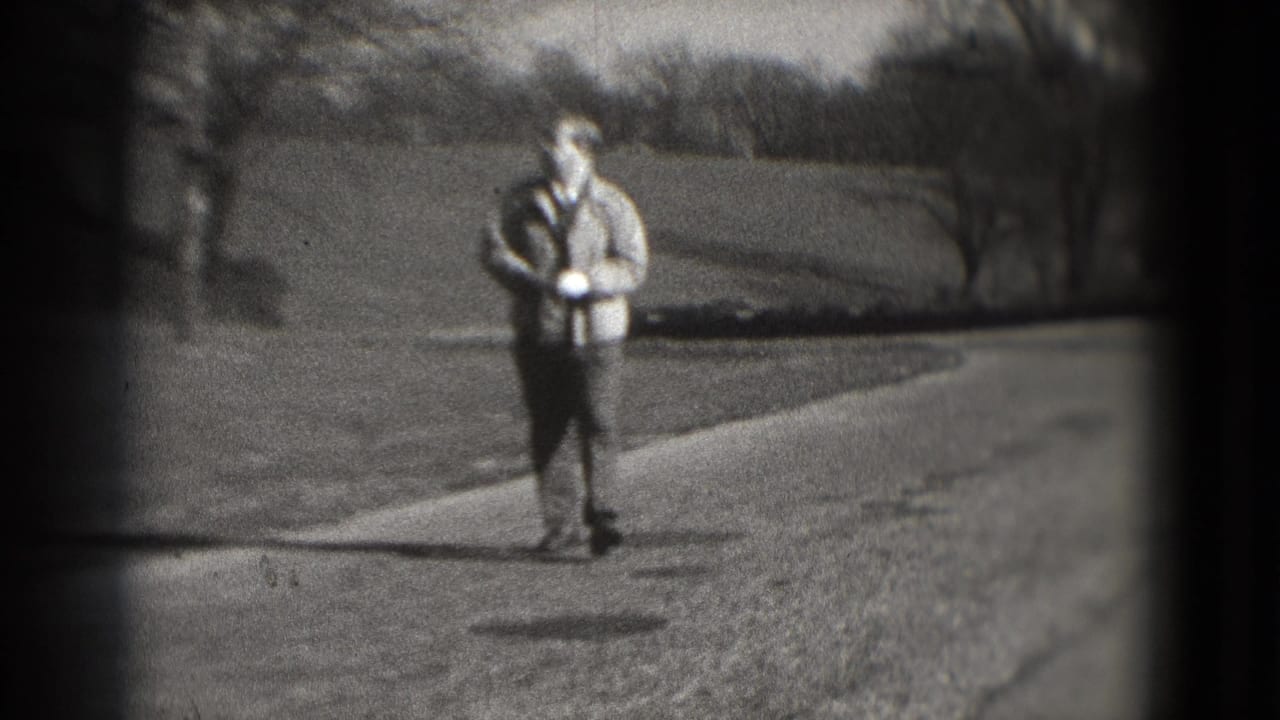 The man walks closer to camera on the paved path, with one hand near a white glove and winter-bare trees in the background.