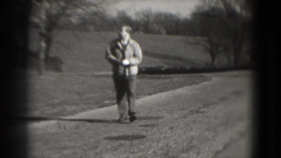 The man walks closer to camera on the paved path, with one hand near a white glove and winter-bare trees in the background.