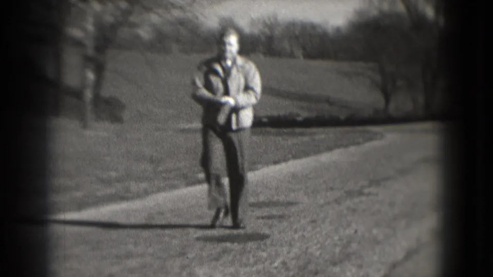 In the 1975 Washington, DC home-movie frame, the man appears to put on or adjust a white glove while walking outdoors.