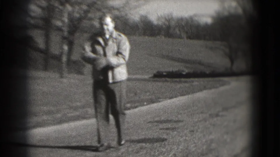 The man stands or slows near the camera, wearing a light jacket and dark pants against a park-like Washington, DC background.