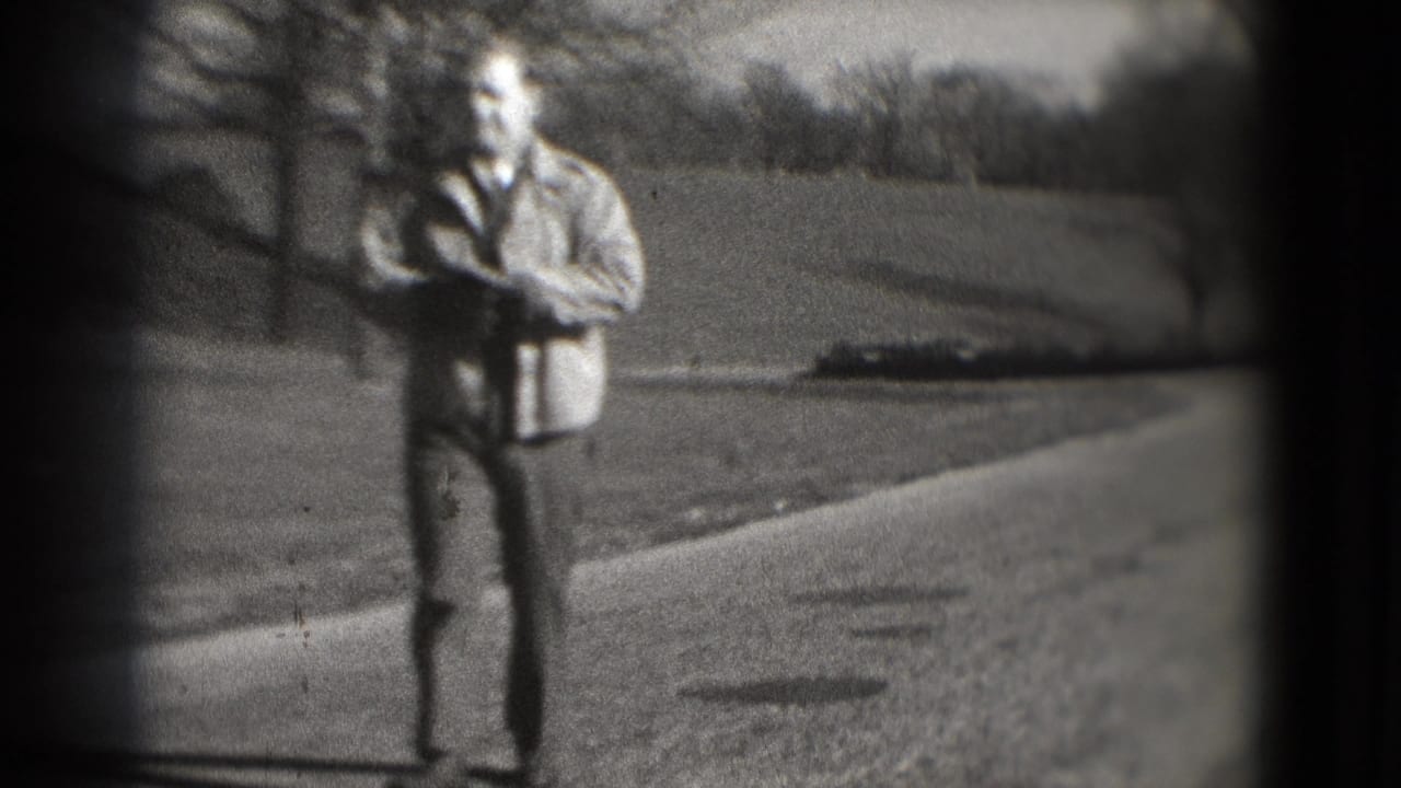 Man in a light jacket walks along a paved park path with grass and bare trees behind him.