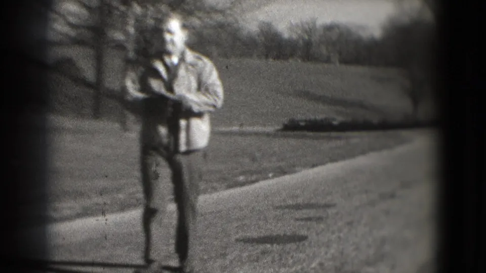 In 1975 Washington, DC home-movie footage, a man walks toward the camera on a paved path bordered by lawn and leafless trees.