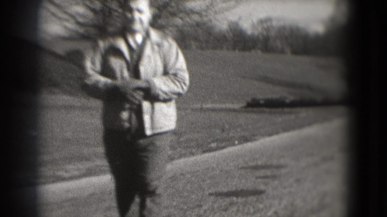The man moves nearer on the park path in grainy 8mm footage, with open lawn, bare trees, and a dark vignette around the frame.