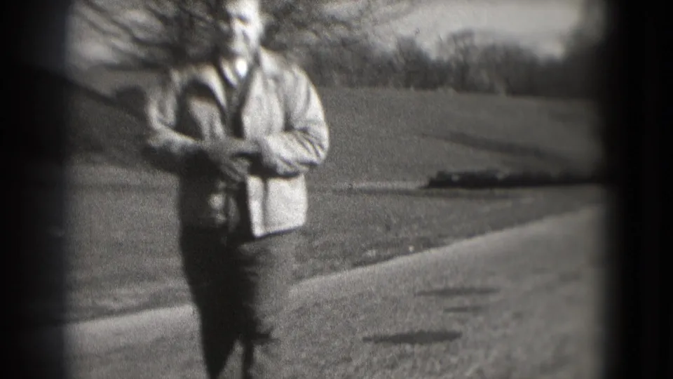 The man moves nearer on the park path in grainy 8mm footage, with open lawn, bare trees, and a dark vignette around the frame.