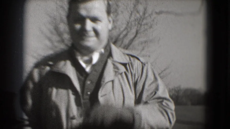 The man smiles near the camera in a soft black-and-white 8mm frame from Washington, DC, with leafless trees and lawn behind him.