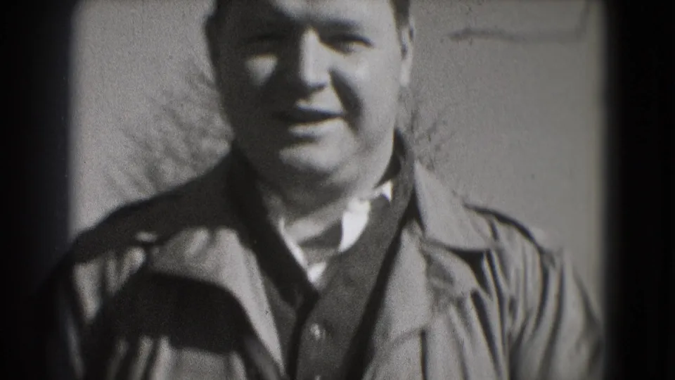 In metadata-supported 1975 Washington, DC footage, a man in a collared jacket and sweater smiles at the camera outdoors.