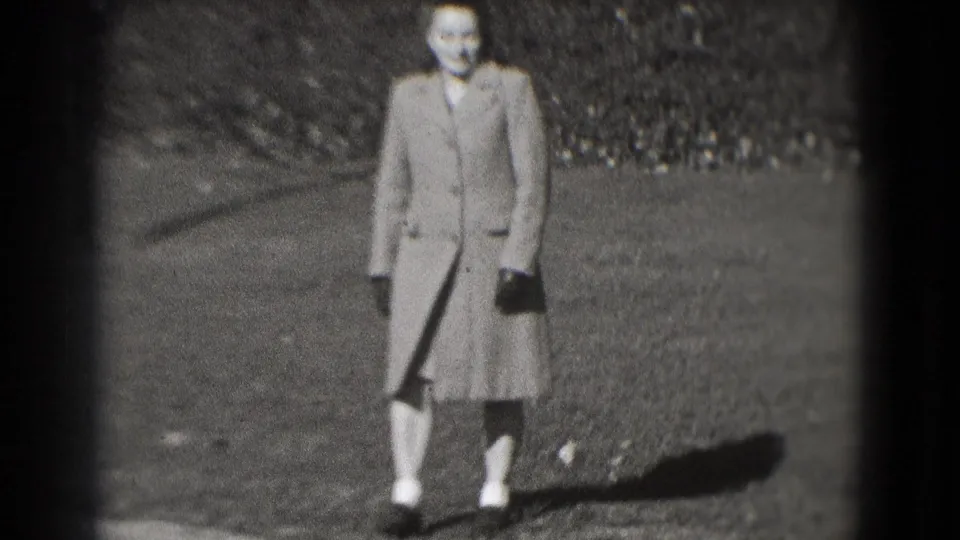 A woman in a long coat and gloves walks on grass in the Washington, DC home-movie footage, slightly blurred by motion and focus.