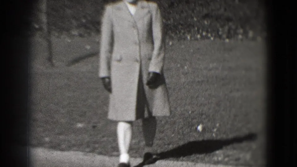The woman continues walking outdoors in a long coat, her upper body partly cropped within the soft 8mm frame.