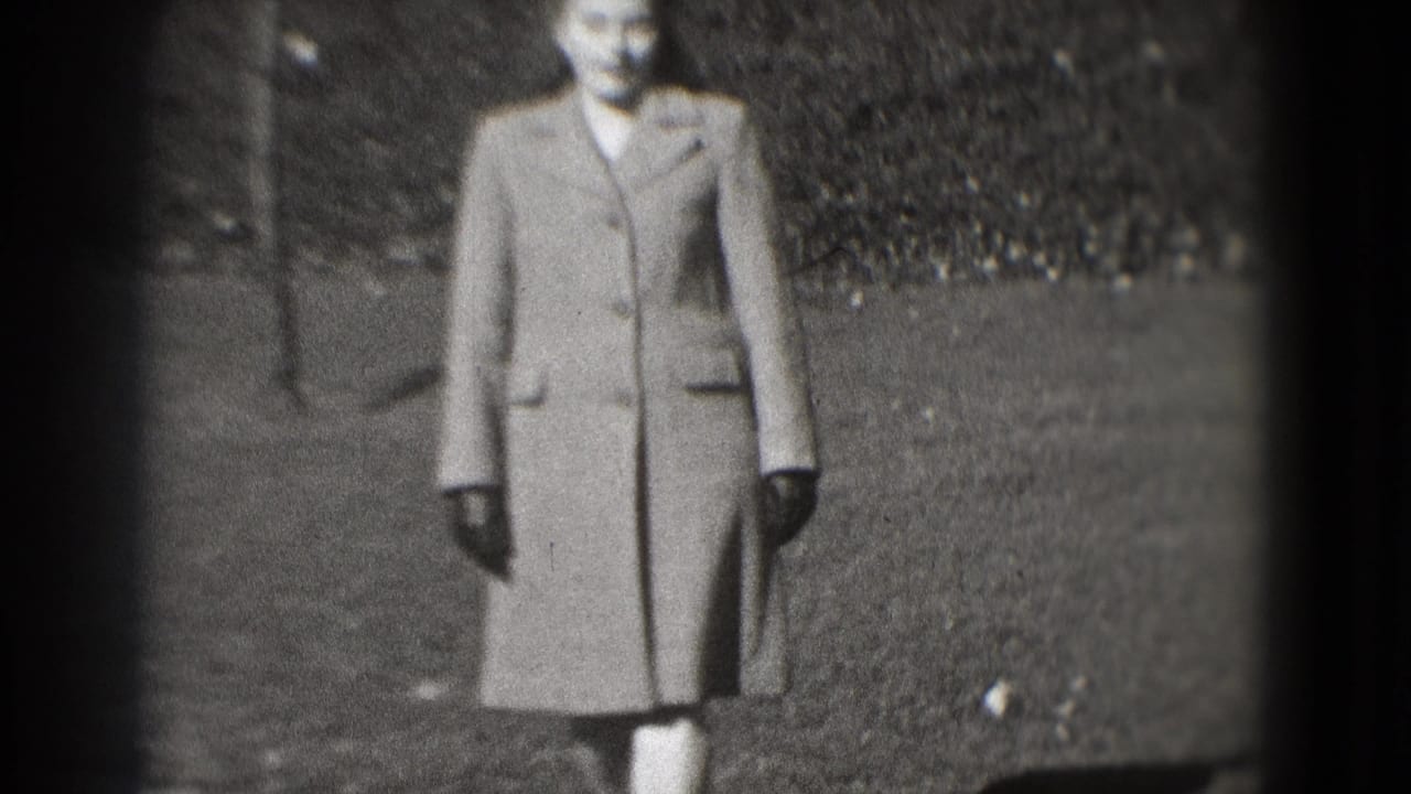 In Washington, DC home-movie footage from 1975, a woman in a tailored coat and gloves stands on a park path near a hedge.