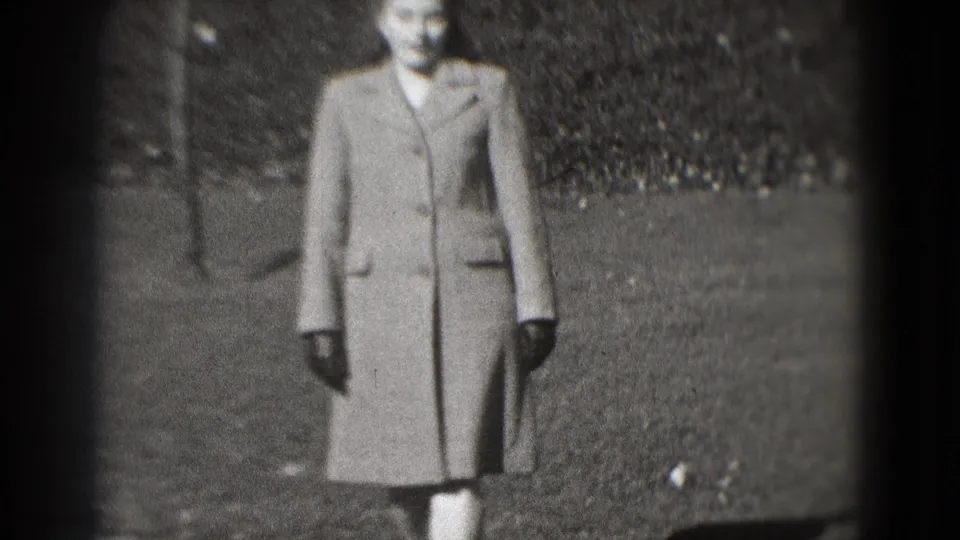 In Washington, DC home-movie footage from 1975, a woman in a tailored coat and gloves stands on a park path near a hedge.