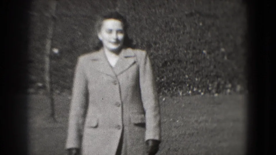 A smiling woman in a tailored coat walks toward the camera in a Washington, DC park setting, filmed on 8mm in 1975.