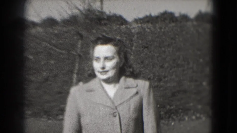 The woman appears closer to the camera, her coat and hairstyle more visible against a hedge and park path in 1975 Washington, DC footage.