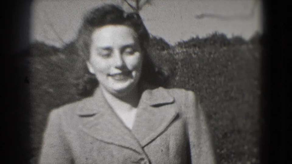 A close 8mm frame shows the woman smiling outdoors in a tailored coat, with a hedge and park path behind her in Washington, DC.
