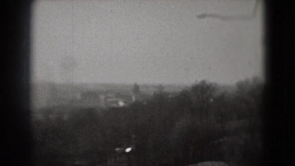 Hazy outdoor city view in black-and-white 8mm, with trees and indistinct buildings visible in the distance.