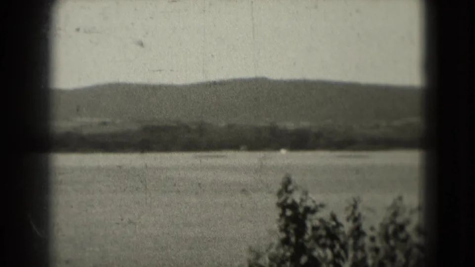 1975 8mm Washington, DC area footage showing calm water, a distant wooded shoreline, low hills, pale sky, and foreground foliage.