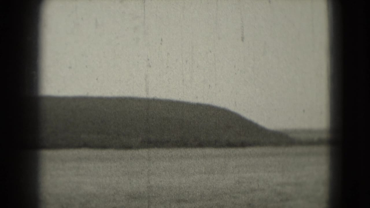 1975 8mm home-movie frame showing calm water and a low distant landmass, with dark film-gate edges and visible grain.
