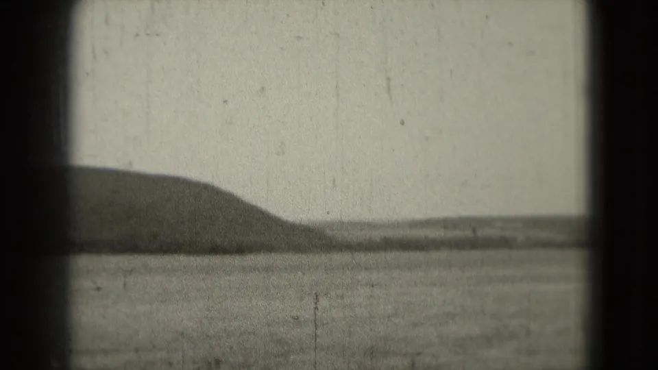 Grainy 1975 8mm view of a low hill or shoreline across still water, partially bordered by dark gate edges.