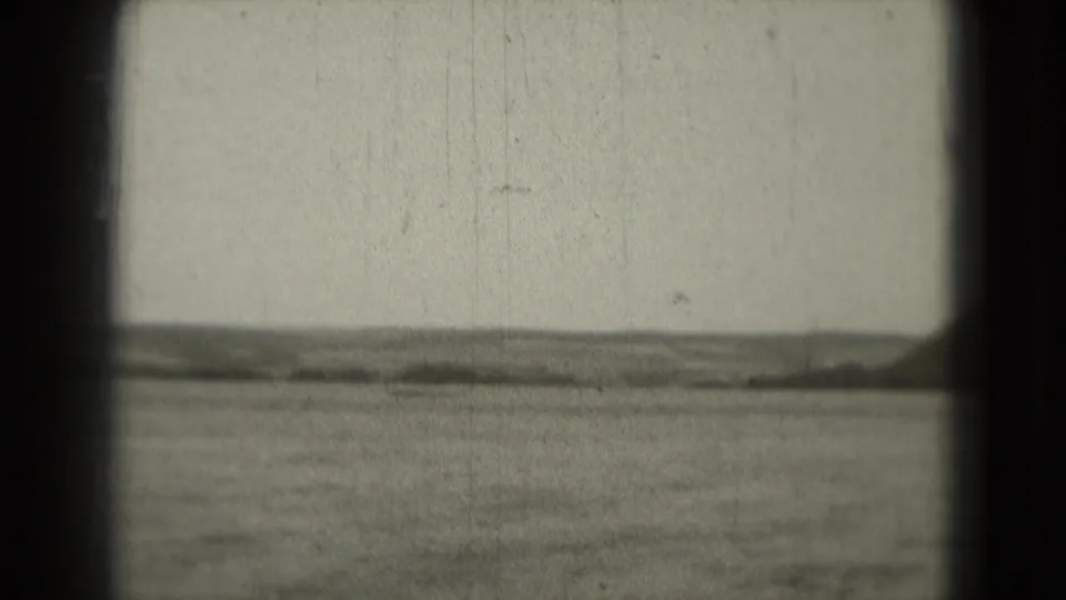 1975 8mm home-movie frame showing open water, hazy shoreline, low hills, and dark gate edges.