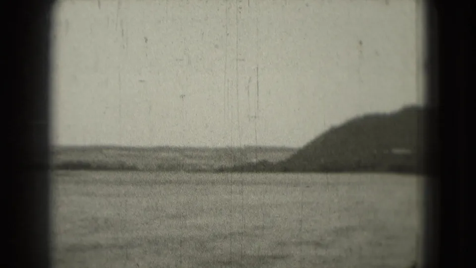 1975 8mm frame with open water, a low shoreline, hazy sky, and a dark hill entering from the right.