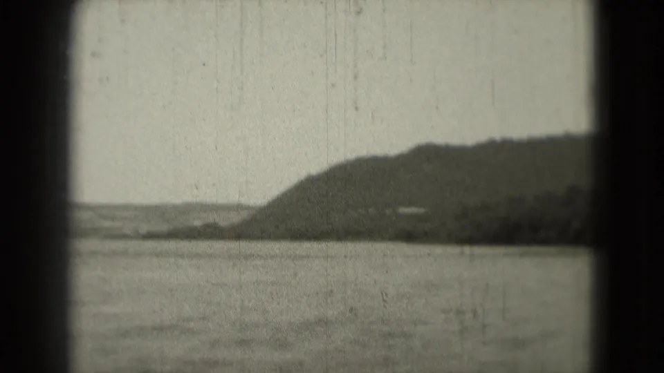 1975 8mm frame showing water in the foreground, a rounded hillside, shoreline structures, and visible film wear.