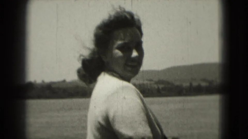 1975 8mm home-movie frame of a windblown woman in a light sweater smiling near open water and distant hills.