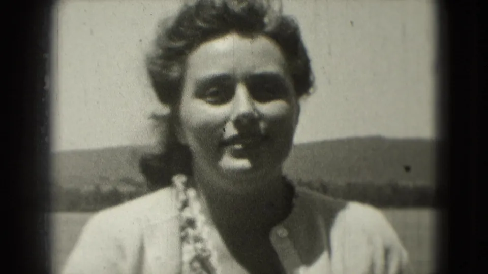 1975 home-movie frame showing a woman facing the camera outdoors, softly focused against bright sky and distant hills.
