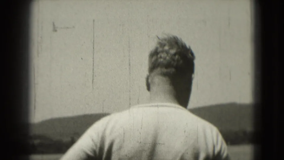 1975 8mm frame of a man viewed from behind outdoors, standing before a bright sky and low distant hills.