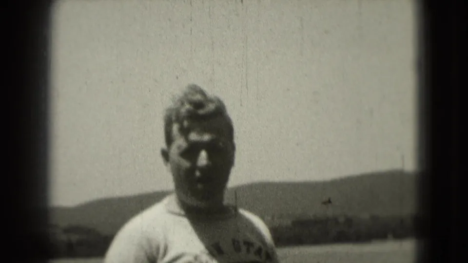 1975 home-movie frame of a man in a T-shirt facing the camera outdoors, with low hills and bright sky behind him.
