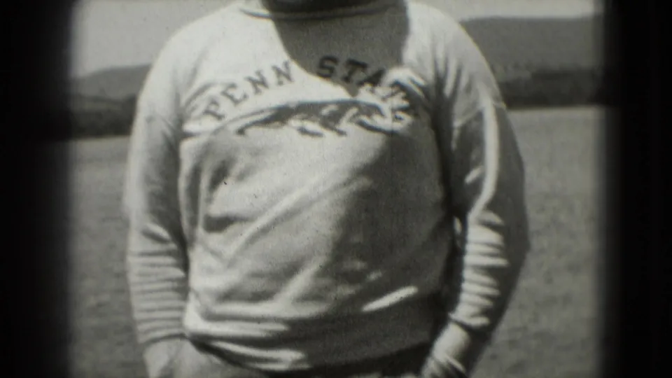 1975 Washington, DC frame showing the man's Penn State sweatshirt and hands near his pockets with water or open background behind him.