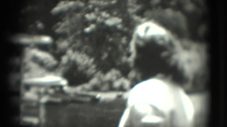 A woman in a light top stands with her back to the camera in 1975 Washington, DC footage, facing a shaded enclosure area.