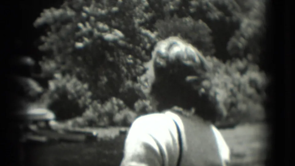 Rear view of a woman in a light top facing trees and an enclosure-like area in 1975 Washington, DC 8mm footage.