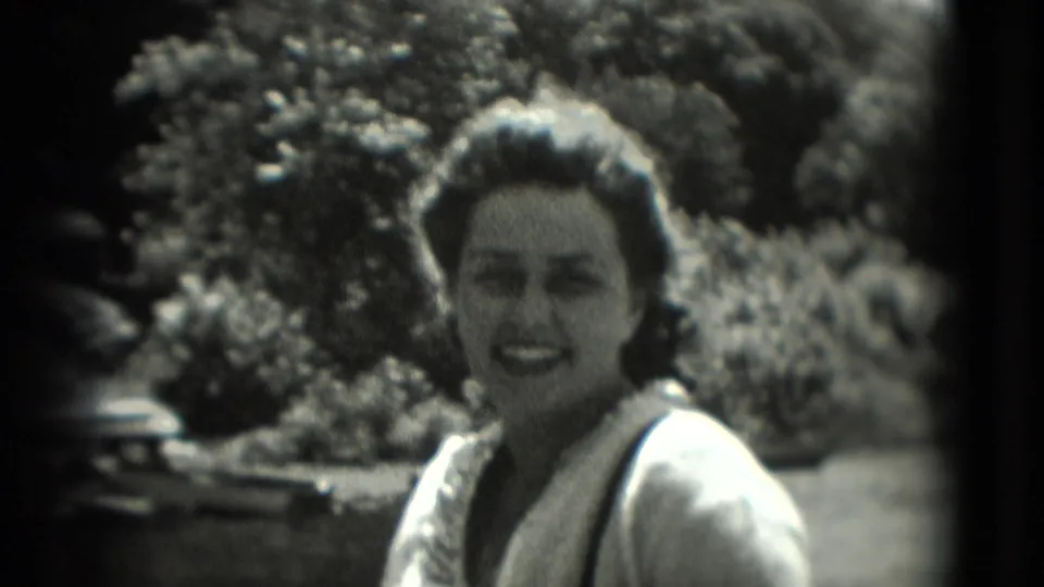 A woman smiles directly toward the camera in 1975 Washington, DC home-movie footage, with foliage and strong daylight behind her.