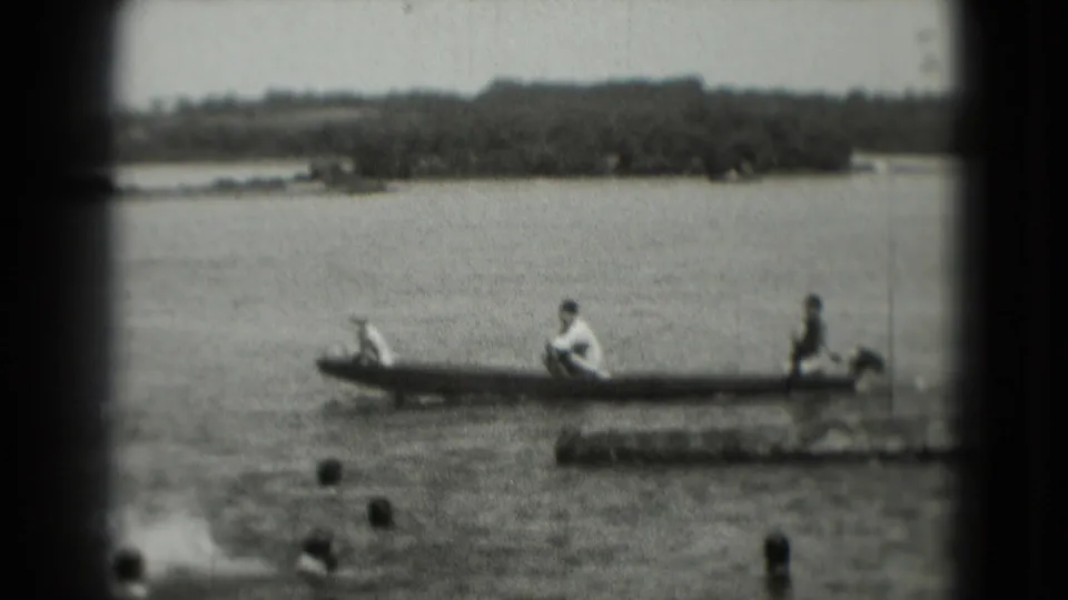Several people sit in a long boat on open water while swimmers are visible nearby, with shoreline and trees in the distance.