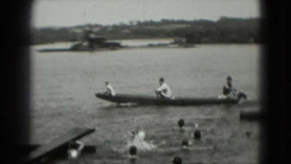 A long boat with people aboard crosses open water near several swimmers, with a tree-lined shore visible in the distance.