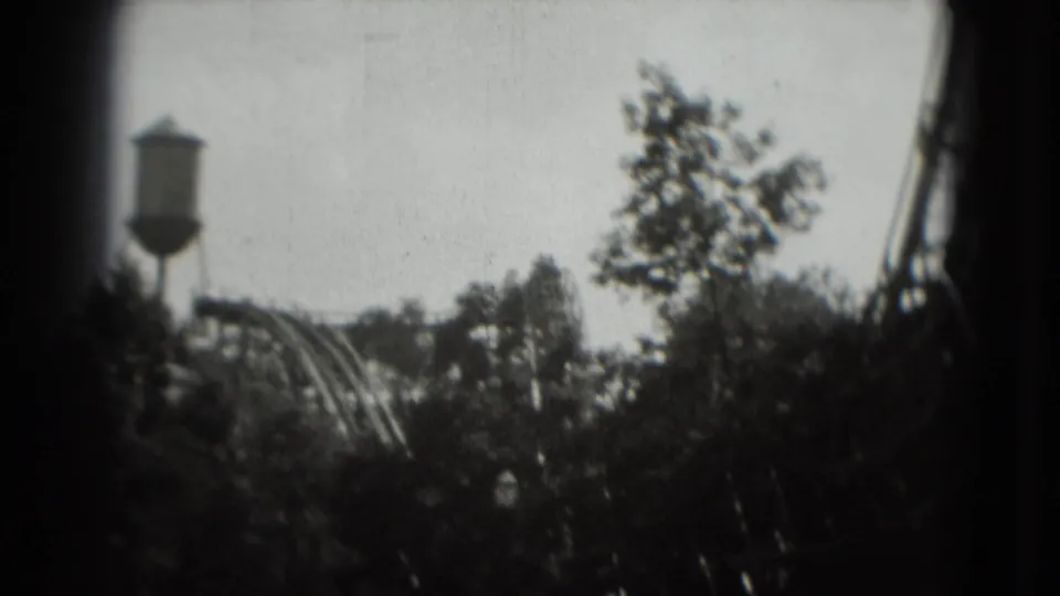1975 8mm Washington, DC view of a water tower and roller coaster seen through a leafy treeline.