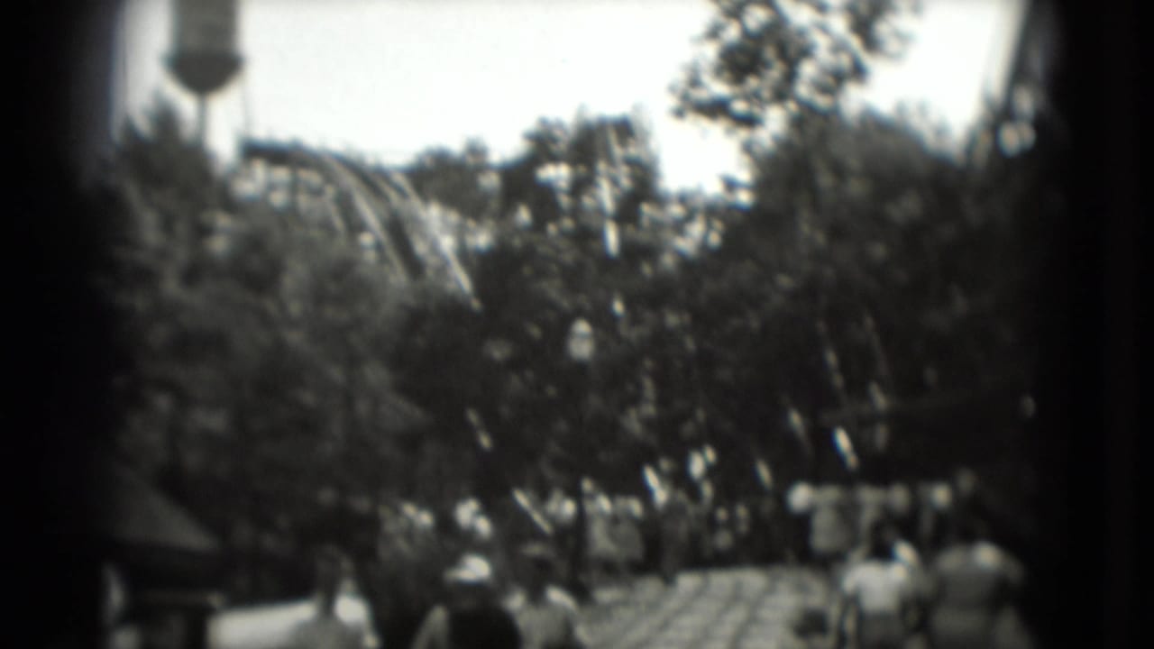 1975 Washington DC home-movie frame showing a wooden roller coaster area, trees, scattered park visitors, and a distant water tower-like form.