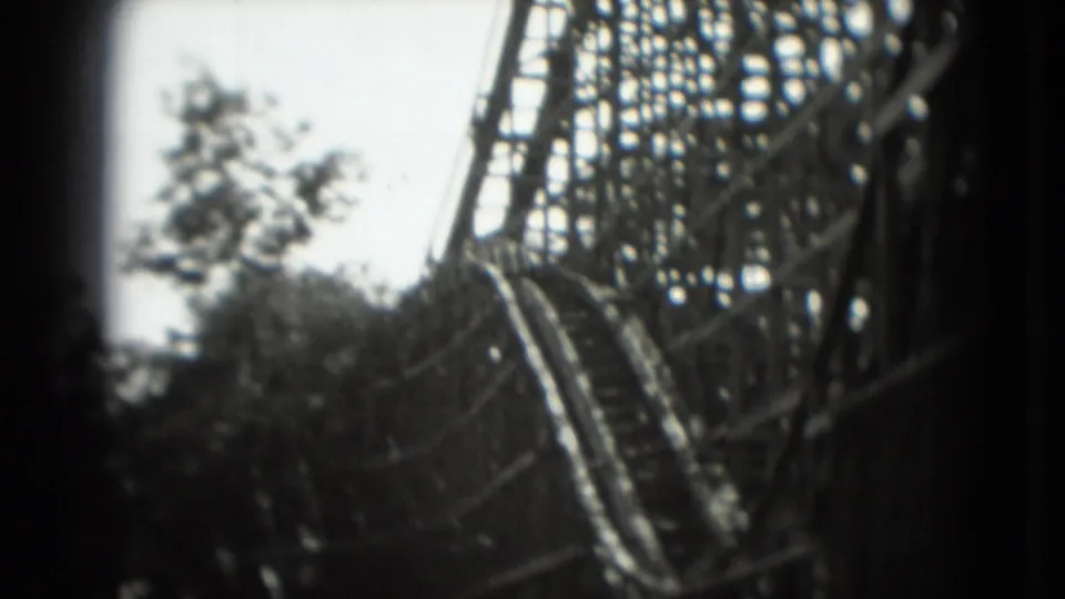 1975 8mm frame showing a closer view of the wooden roller coaster track and support lattice beside trees in the amusement park.