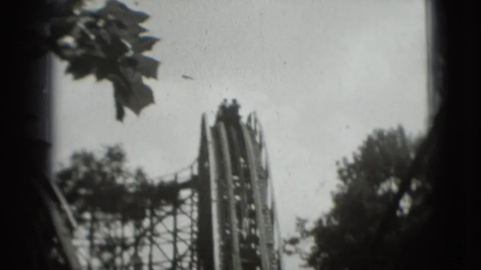 1975 8mm Washington, DC area rollercoaster view with indistinct riders near the lift hill top and trees around the track.