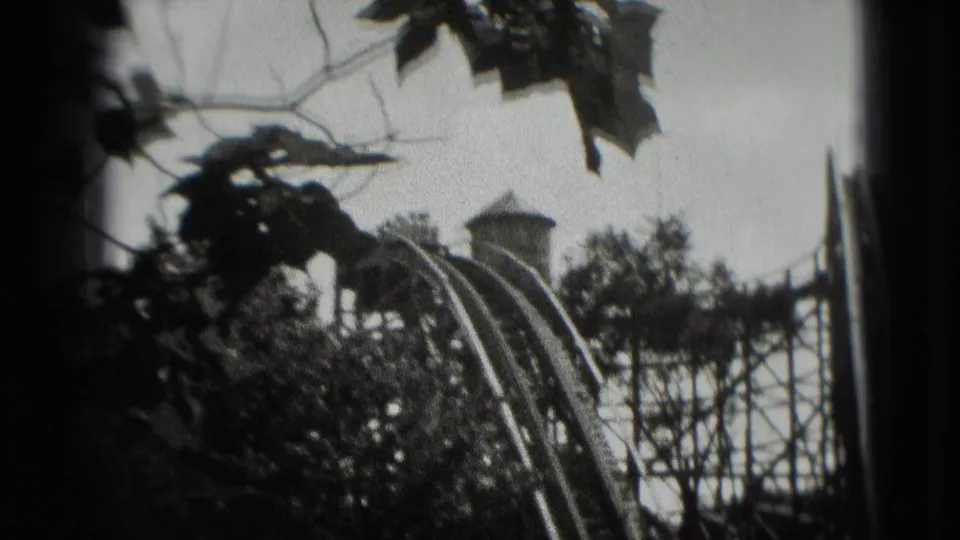 1975 8mm home-movie frame showing a wooden roller coaster structure partly obscured by trees, with a train near the track.
