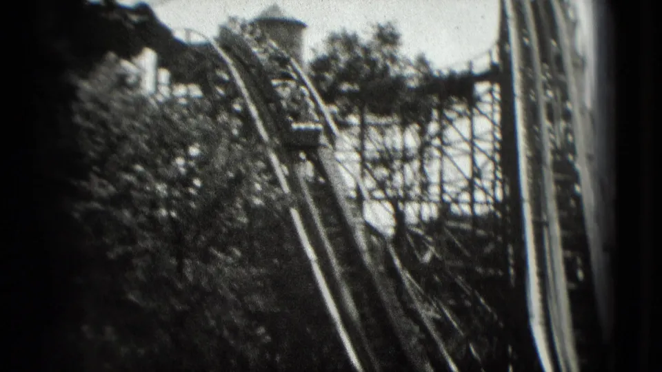 Grainy 1975 8mm frame with a roller coaster train riding along elevated track near trees and timber support structure.