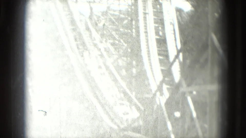 Brightly exposed 8mm frame showing portions of a roller coaster track and support structure, softened by glare and film grain.