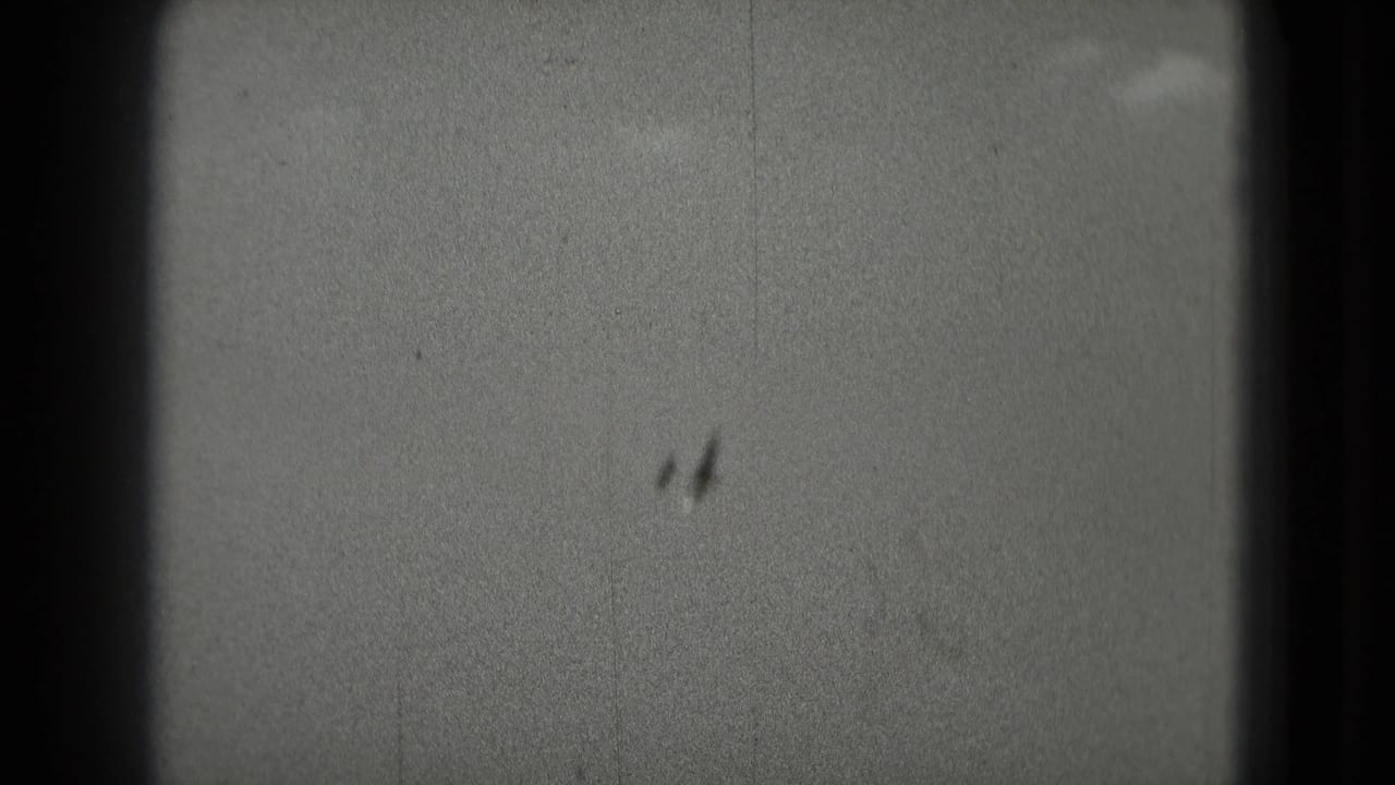 Two distant aircraft-like silhouettes pass through a grainy cloudy sky in 1975 8mm home-movie footage.