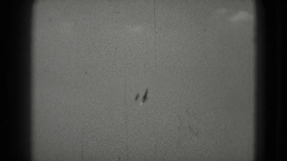 Two distant aircraft-like silhouettes pass through a grainy cloudy sky in 1975 8mm home-movie footage.