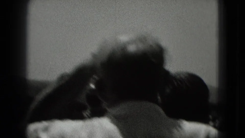 A handheld 8mm frame shows blurred onlookers from behind, gathered outdoors under a cloudy sky.