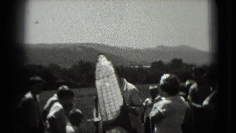 People gather outdoors in a rural-looking area with low hills beyond, in grainy 1975 8mm footage linked to Washington, DC metadata.