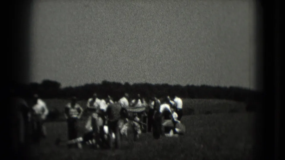 1975 Washington DC home-movie frame showing a small crowd standing and crouching in a grassy field under an overcast sky.