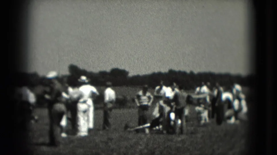 1975 Washington DC home-movie frame with adults in summer clothing gathered across a grassy field, some turned toward an off-camera event.